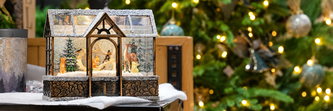 Maison décorative de Noël illuminée avec animaux de la forêt, devant un sapin aux lumières dorées.